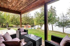 An outdoor patio with wicker furniture and a glass table, overlooking a lake and a grassy area with trees. - Stunning European waterfront escape with hot tub (Lyndhurst)