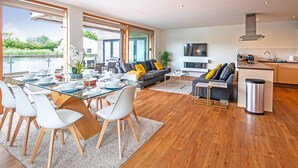 A modern living room with a dining area, a grey sofa with yellow cushions, a TV mounted on the wall, and a kitchen area with a stainless steel trash bin. - Waters Edge 04, Waterside Lodge (P) - Lakeside Luxury House, Hot Tub, Sleeps 8 (Cirencester)