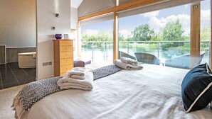 A bedroom with a large bed, a wooden dresser, a bathroom, and a balcony with a view of trees and a body of water. - Waters Edge 04, Waterside Lodge (P) - Lakeside Luxury House, Hot Tub, Sleeps 8 (Cirencester)