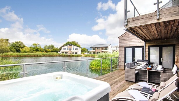 A hot tub on a wooden deck with outdoor furniture, overlooking a body of water and buildings in the background. - Waters Edge 04, Waterside Lodge (P) - Lakeside Luxury House, Hot Tub, Sleeps 8 (Cirencester)