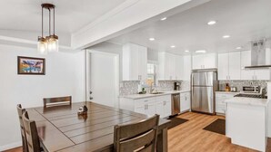 A modern kitchen with white cabinets, a stainless steel refrigerator, and a dining area with a wooden table and chairs. - Modern and Clean with a Spa! Close to Snow Summit. (Big Bear)