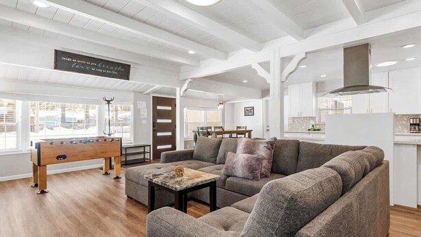 A spacious living room with a sofa, a foosball table, and a coffee table. There is a kitchen area with white cabinets and a hanging sign. - Modern and Clean with a Spa! Close to Snow Summit. (Big Bear)