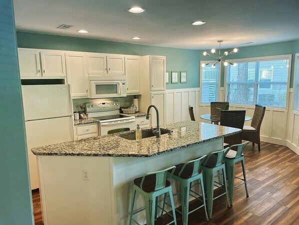 A kitchen with white cabinets, a granite countertop, a built-in microwave, and a sink. There are three bar stools and a dining table with chairs. - 2BR/2BA w/ Rainfall Showers, Screened Porch, 1st Floor, Pool, Gated Community (Saint James)