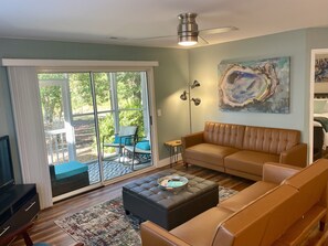 A living room with a large sliding glass door leading to a patio, a brown leather sofa, a black ottoman, and a floor lamp. - 2BR/2BA w/ Rainfall Showers, Screened Porch, 1st Floor, Pool, Gated Community (Saint James)
