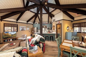 A spacious living area with a high ceiling, wooden beams, a television, a fireplace, and a kitchen in the background.