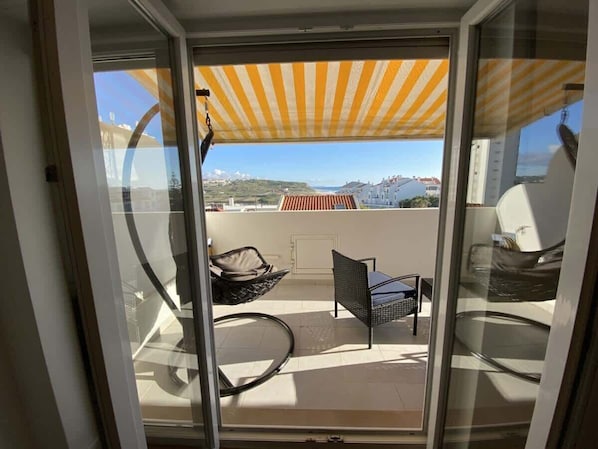 A balcony with a yellow-striped awning, a wicker chair, a metal chair, and a view of buildings and hills.