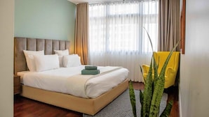 Quarto de hotel com uma cama grande, cabeceira, janela com cortinas, uma cadeira amarela e uma planta.