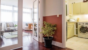 Uma sala de estar e jantar integrada, com uma planta em vaso, uma máquina de lavar e uma cozinha com armários.