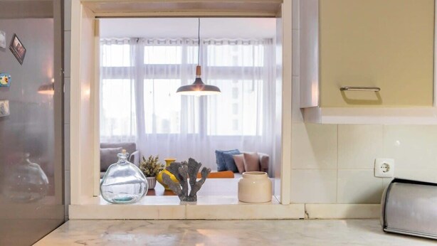 Uma cozinha com bancada de mármore, uma luminária pendente e vista para uma sala de estar com sofá e cortinas.
