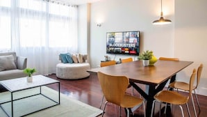 Uma sala de estar moderna com uma mesa de jantar de madeira, assentos confortáveis, uma TV de tela plana e uma grande janela com cortinas translúcidas.