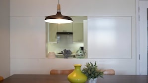 Uma mesa de jantar com um vaso amarelo e uma planta em vaso, uma luminária pendente e uma cozinha ao fundo.