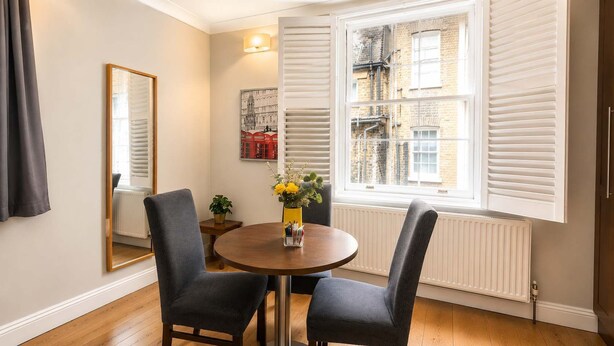 Un coin repas avec une table ronde, deux chaises, un vase de fleurs, un miroir et une fenêtre avec des stores.