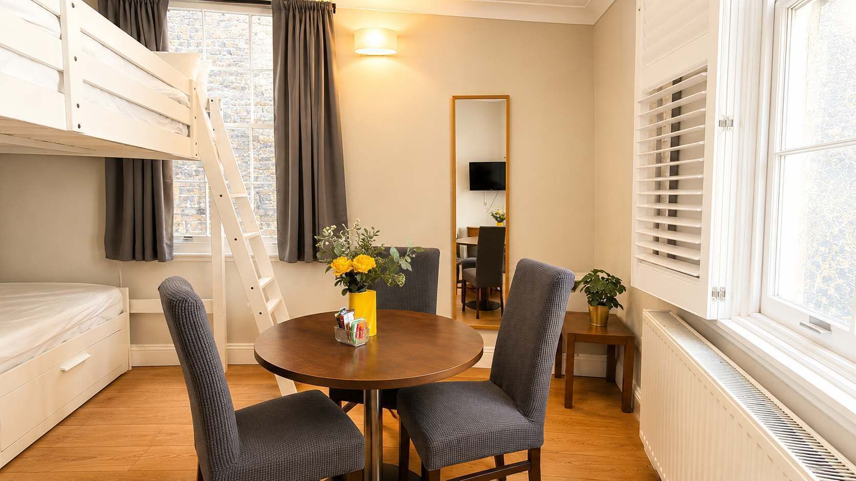 Une pièce avec un lit superposé, une table de salle à manger ronde avec des chaises, une petite table avec une plante, un miroir et un radiateur.