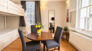 Une pièce de taille modeste comprenant un lit superposé, une table à manger avec des chaises, une petite table ornée d’un vase de fleurs et une fenêtre avec des stores.