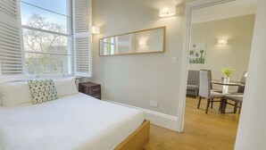 Een slaapkamer met een bed, nachtkastje en een raam met zonwering. Door een geopende deur is een eethoek met stoelen en een tafel zichtbaar.