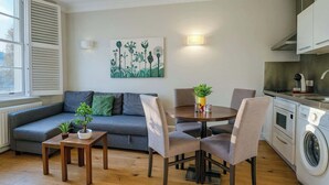 Een ruimtelijke leefruimte met een bank, een eettafel, stoelen, een kitchenette met een wasmachine, en een raam met zonwering.