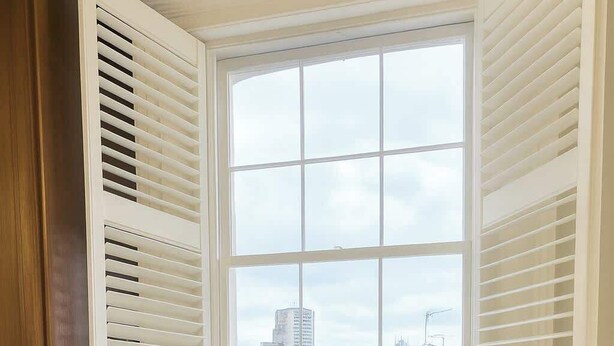 Ein Eckzimmer mit Erkerfenster, zwei Sessel, einem kleinen Tisch und einer gelben Vase mit Blumen.