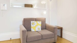 Ein modernes Wohnzimmer mit einem beige-farbenen Sofa, einem dekorativen Kissen mit Margeritenmuster, einem kleinen Holztisch und einem wandmontierten Spiegel.