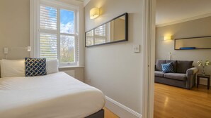 Een slaapkamer met een groot bed, een raam met jaloezieën, een spiegel, en een bank in de aangrenzende kamer.