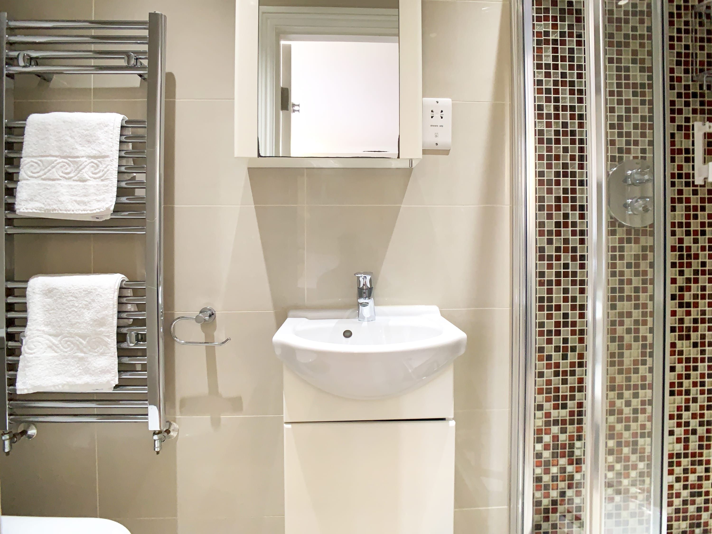 A bathroom with a white sink, a towel rack with two towels, a mirror, and a shower with mosaic tiles.