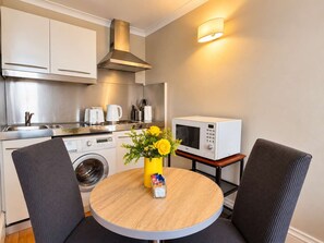 Een functionele keuken met een magnetron, broodrooster en wasmachine, een ronde houten tafel met stoelen, en een vaas met gele bloemen.