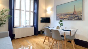 A room with a wooden floor, a dining table with chairs, a potted plant, a flat-screen TV, and a painting of a cityscape. - S3 - Ground floor apartment by Marylebone (London)