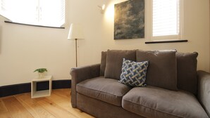 A living room with a brown sofa, a small white side table with a plant, and a floor lamp. - S4 - Comfy Loft Apartment 10 Mins to Oxford Street (London)
