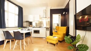 A modern kitchen with white cabinets, a dining table with black chairs, a yellow armchair, and a large TV screen displaying the Netflix logo. - S4 - Comfy Loft Apartment 10 Mins to Oxford Street (London)