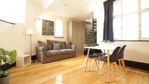 A modern living room with a wooden floor, a grey sofa, a dining table with chairs, and a staircase in the background. - S4 - Comfy Loft Apartment 10 Mins to Oxford Street (London)