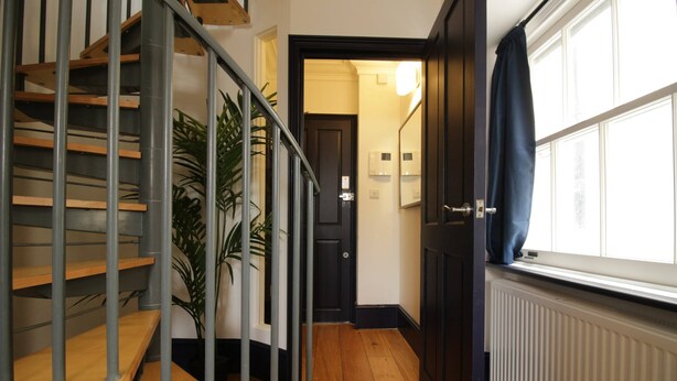 A staircase with wooden steps and metal railings, leading to a door with a light switch and a wall-mounted light fixture. - S4 - Comfy Loft Apartment 10 Mins to Oxford Street (London)