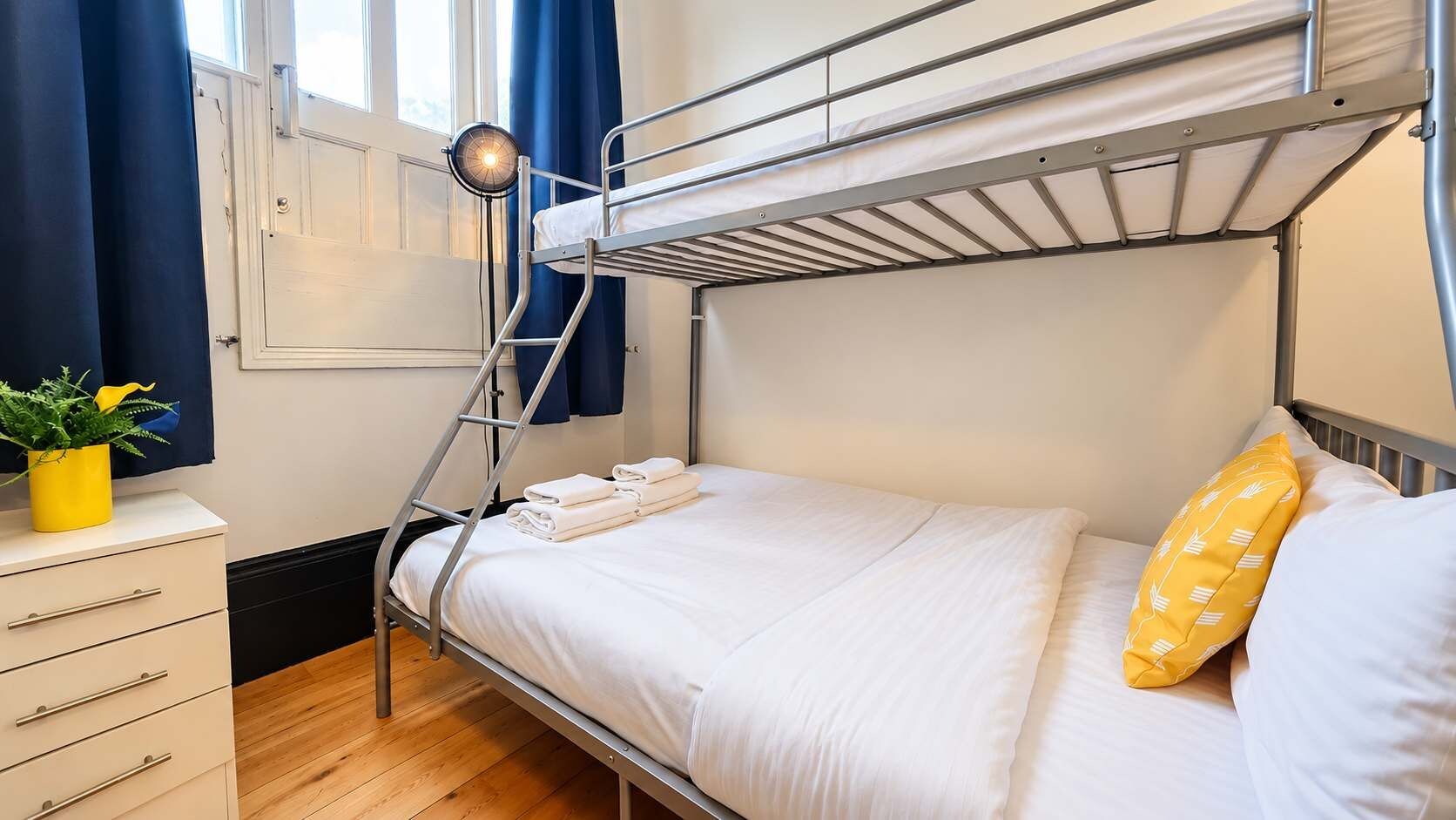 Una cama litera con escalera, una mesita de noche con una planta en maceta y una ventana con cortinas.