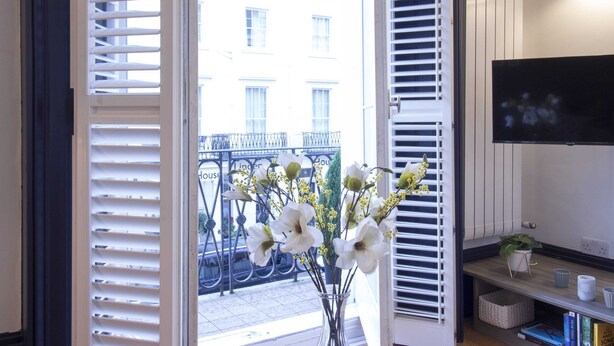 A room with a large window, white shutters, a potted plant, a flat-screen TV, and a bookshelf. - S5 - Cute Apartment with Balcony by Oxford Street (London)