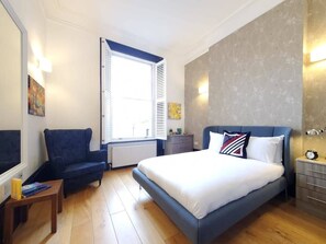 Ein Schlafzimmer mit einem Bett, einem Sessel, einem Nachttisch, einer Kommode und einem Fenster mit Rollläden.