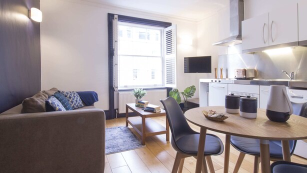 Un espace de vie compact comprenant un canapé, une table à manger avec des chaises, un coin cuisine équipé d’appareils électroménagers et une fenêtre avec des stores.