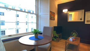 A modern living room with a round table, a chair, a potted plant, and a view of a building through a large window. - S8 - Comfy Apartment in the heart of Hyde Park (London)