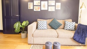 Una sala de estar con un sofá color crema, cojines estampados, una planta en maceta y una pared con cuadros.
