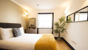 Una cama bien hecha con cabecera, mesita de noche, planta en maceta y ventana con persianas.