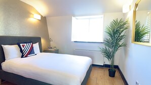 A neatly made bed with white linens, a bedside lamp, and a small table with books and an alarm clock. A large window allows natural light into the room, and a potted plant is placed near the window. - S11 - Attractive Penthouse in Paddington (London)
