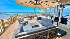 A deck with outdoor seating, including sofas and a table, overlooking a beach and ocean.