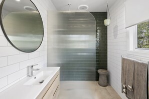 A modern bathroom with a white sink, a round mirror, a shower with a glass door, and a window with a view of greenery.