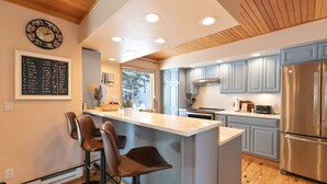 Une cuisine moderne dotée d’un îlot central, de tabourets de bar, d’un plafond en bois et d’un réfrigérateur en acier inoxydable.