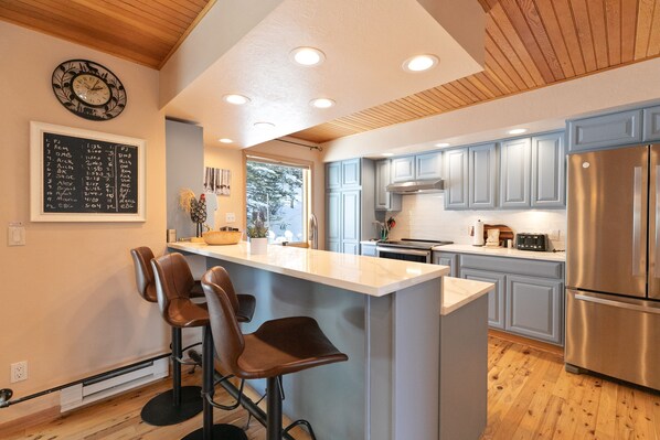 A modern kitchen with a central island, bar stools, wooden ceiling, and a stainless steel refrigerator. - Sunrise Ridge Townhome: Hot Tub, Stunning Views (Breckenridge)