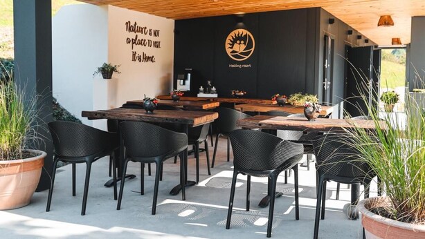 An outdoor seating area with wooden tables and black chairs, a sign with a nature quote, and a logo for 'Nesting Resort'. - Tends Resort Posestvo SONČNI RAJ (Maribor)