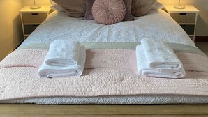 Un lit bien fait, recouvert d’une couverture rose, avec deux oreillers et deux serviettes pliées. Au-dessus du lit, on peut voir un tableau représentant un paysage côtier.