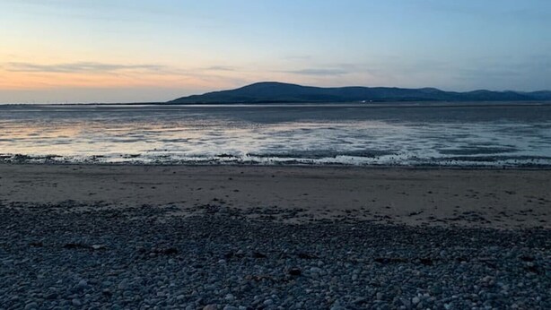 Une plage de galets avec une eau calme et des montagnes qui se dessinent à l’horizon.