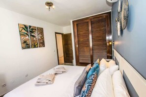 A bedroom with a bed, towels, a headboard, a wall-mounted artwork, and a door with a shutter. - Casa Gecko | Rubicon Retreat by Villas Now (Playa Blanca)