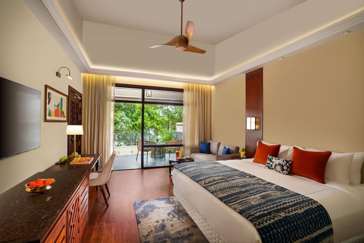 A hotel room with a bed, a desk, a TV, and a balcony with a view of greenery.