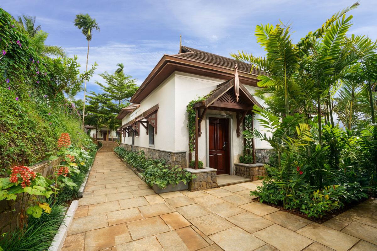 A pathway leading to a house with a stone facade, surrounded by lush greenery and a clear sky.