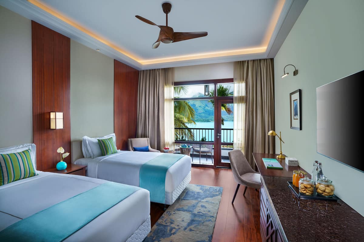 A hotel room with two beds, a desk with a chair, a TV, and a balcony with a view of the sea.