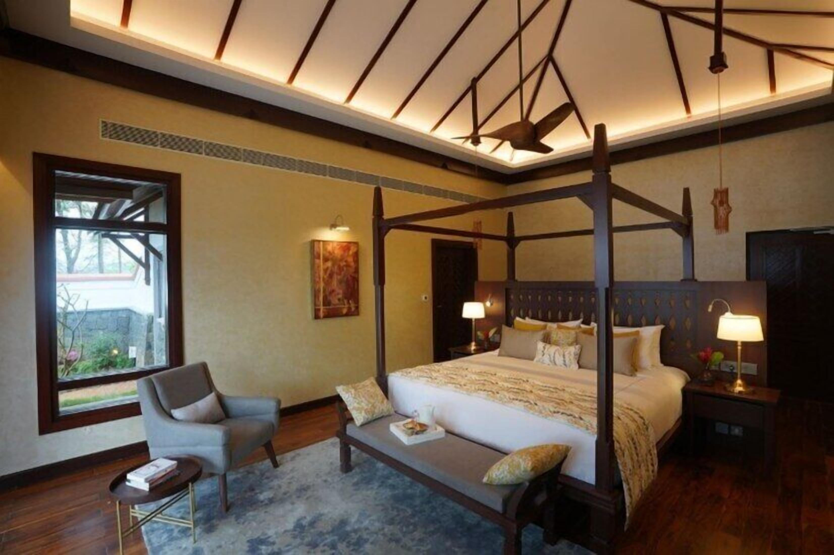 A four-poster bed with a bench, a chair, and a bedside table with a lamp.
